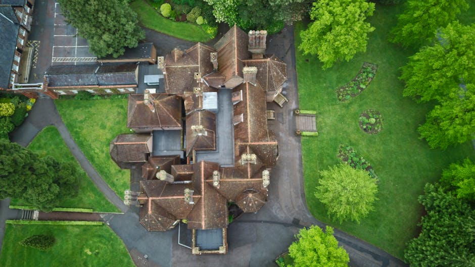 An aerial view of a large, historic house with a complex roof structure covered in reddish-brown tiles, surrounded by lush green trees and well-maintained gardens. The property features multiple chimn