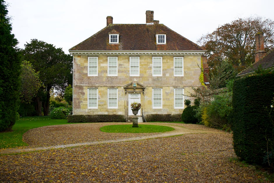 An aerial view of a large, historic house with a complex roof structure covered in reddish-brown tiles, surrounded by lush green trees and well-maintained gardens. The property features multiple chimn
